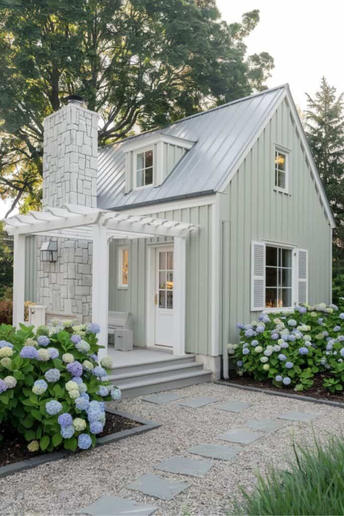 Sage-green cottage with white trim, stone chimney, and blooming hydrangeas